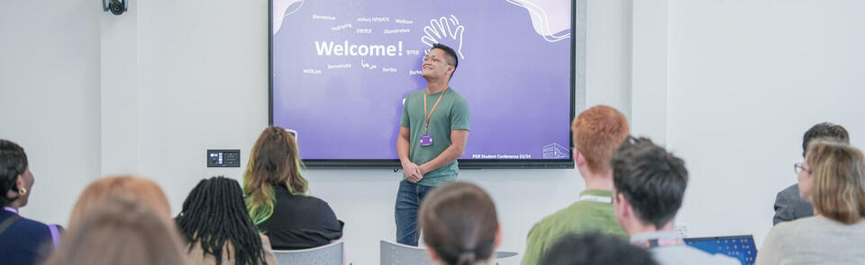 Photo of students in a lecture theatre looking towards a speaker in front of a smart board. The smartboard says 'Welcome'